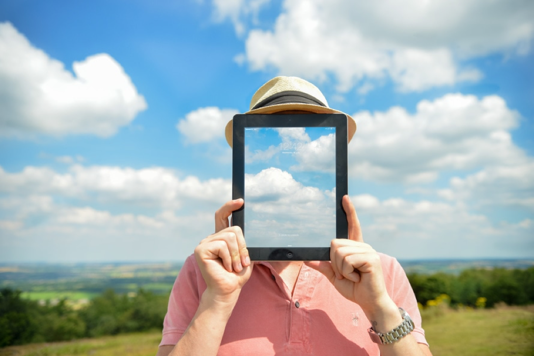 A person holding a tablet in front of their face, with a cloud image that matches the sky in the background.